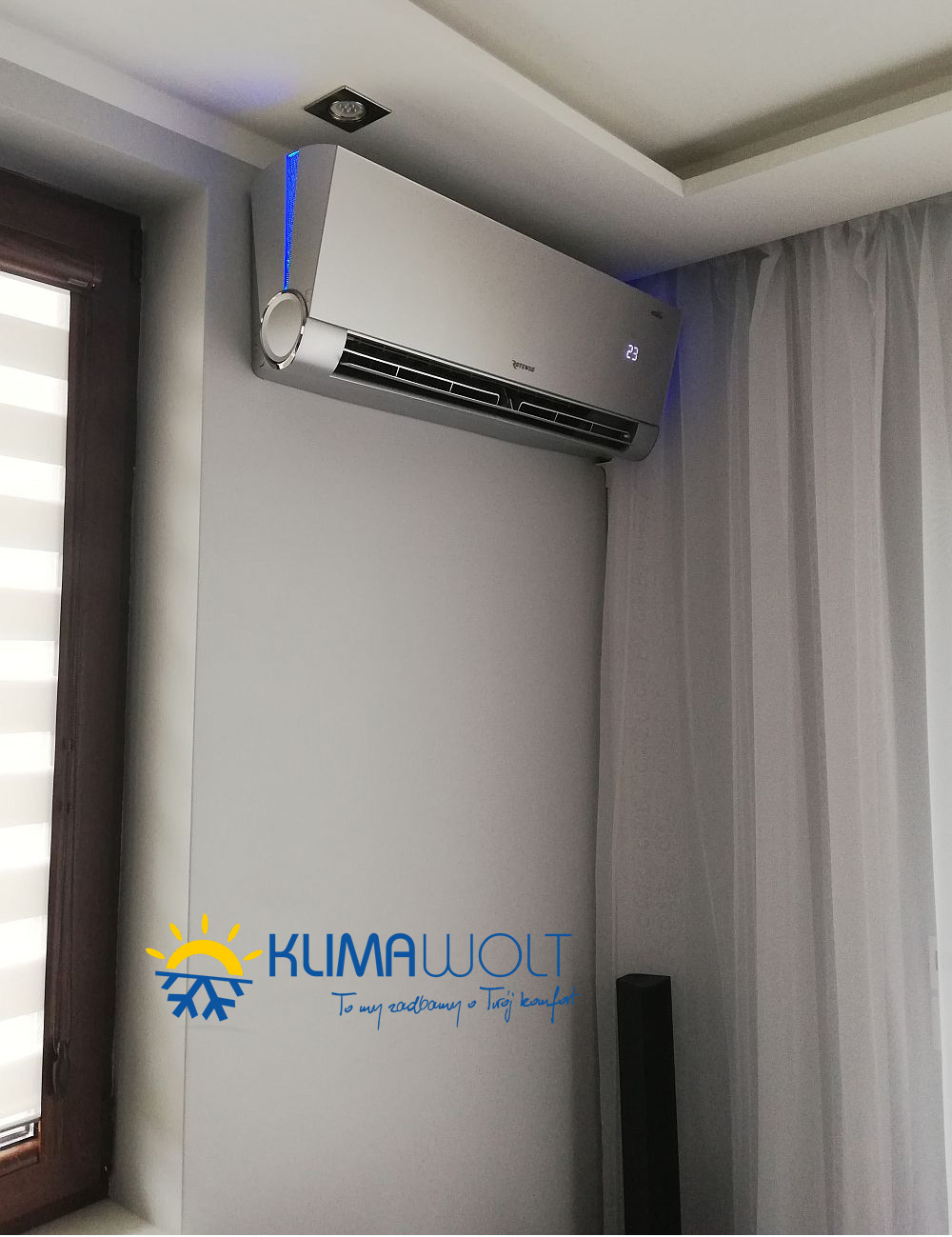 Nowoczesny, srebrny klimatyzator ścienny z niebieskim podświetleniem LED, zamontowany w jasnym pomieszczeniu z zasłonami i logo firmy na ścianie.