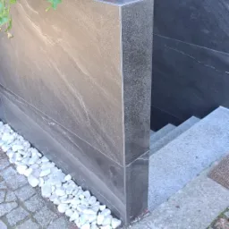 Zewnętrzna ściana budynku wykończona ciemnoszarą płytą imitującą kamień, z donicą na górze i białymi kamykami u podstawy, prowadząca do schodów.