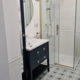 Nowoczesna łazienka z kabiną prysznicową w złotych ramach, umywalką na szafce w kolorze granatowym, okrągłym lustrem i wzorzystą podłogą.