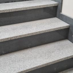 Oskar Hadrian - Wykończone schody wewnętrzne z szarego granitu, z ciemnymi podstopnicami i listwami przy ścianie, na dolnym stopniu zwinięty kabel.