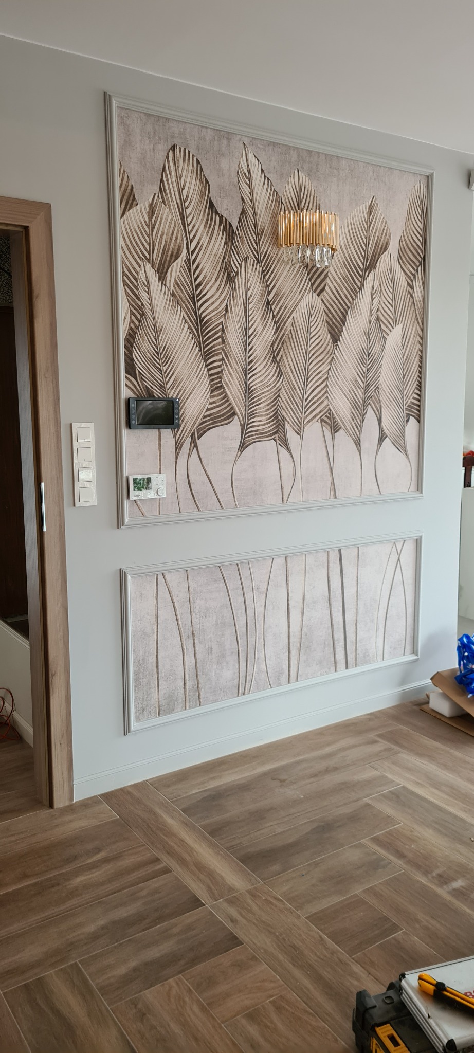 Ściana w Chwałowicach z dekoracyjnymi panelami z motywem liści i abstrakcyjnymi pionowymi liniami, oświetlona złotym kinkietem, z drewnopodobną podłogą ułożoną w jodełkę.