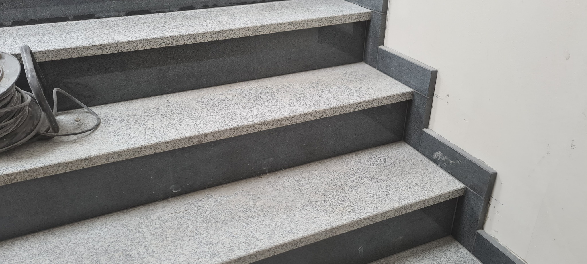 Wykończone schody wewnętrzne z szarego granitu, z ciemnymi podstopnicami i listwami przy ścianie, na dolnym stopniu zwinięty kabel.