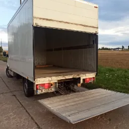 Otwarty biały samochód dostawczy z opuszczoną rampą załadowczą na betonowym placu, w tle zaparkowane samochody osobowe i pole.