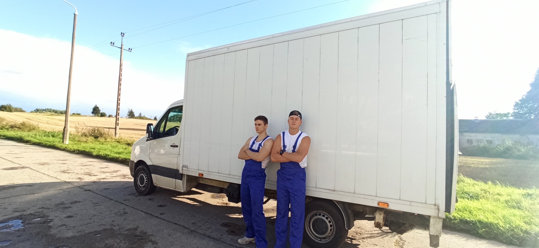 Dwóch młodych mężczyzn w niebieskich kombinezonach, z założonymi rękami, stoi przed białym samochodem dostawczym na tle wiejskiego krajobrazu z polami i słupami energetycznymi.