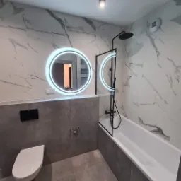 Nowoczesna łazienka z marmurowymi płytkami, okrągłym lustrem z podświetleniem LED, białą wanną z czarną armaturą i szklanym parawanem, oraz szarą zabudową z podwieszaną toaletą.