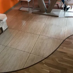 Beżowe płytki podłogowe o fakturze kamienia, ułożone w kwadraty, z widocznym fragmentem drewnianego parkietu i obudowy kominka z dekoracyjnym ułożeniem drewna