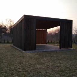 Nowoczesna, minimalistyczna konstrukcja z ciemnego drewna z ażurowymi ścianami, prawdopodobnie altana lub zadaszenie tarasu, na tle trawnika i zachodzącego słońca.