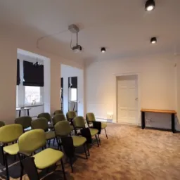 Jasne wnętrze sali konferencyjnej z rzędem zielonych krzeseł, projektorem pod sufitem i flipchartem z zapiskami.