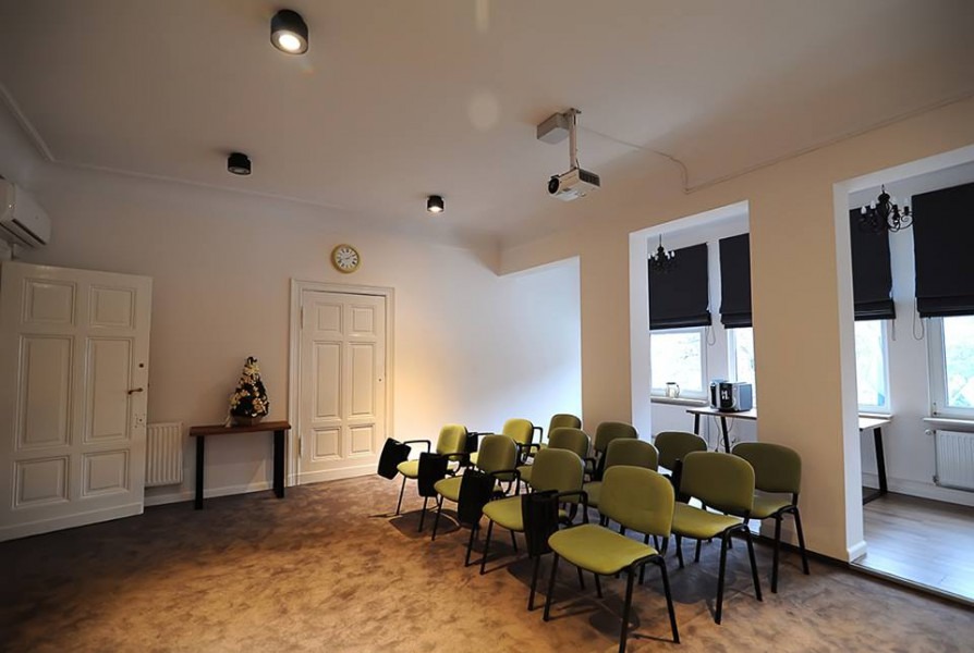 Jasne wnętrze sali konferencyjnej z rzędem zielonych krzeseł, projektorem sufitowym i zegarem ściennym w tle.