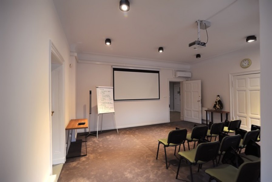 Sala konferencyjna z projektorem, ekranem, flipchartem i rzędami krzeseł, gotowa do przeprowadzenia szkolenia lub prezentacji.