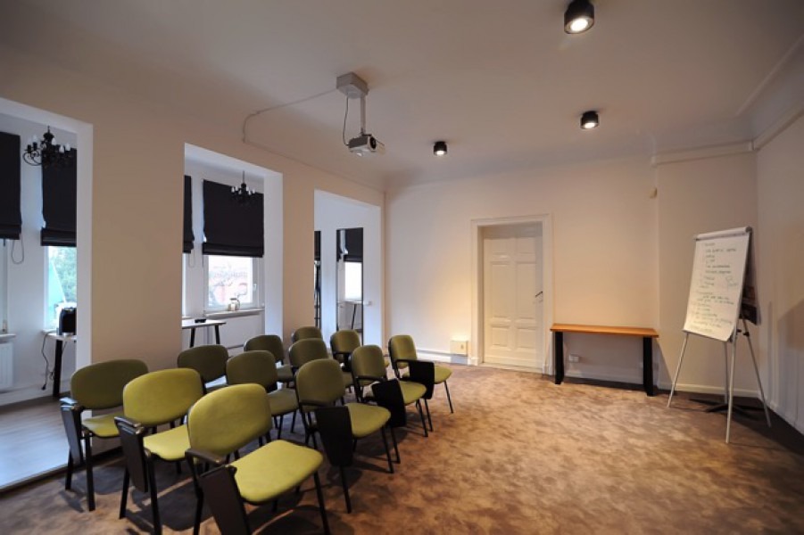 Jasne wnętrze sali konferencyjnej z rzędem zielonych krzeseł, projektorem pod sufitem i flipchartem z zapiskami.