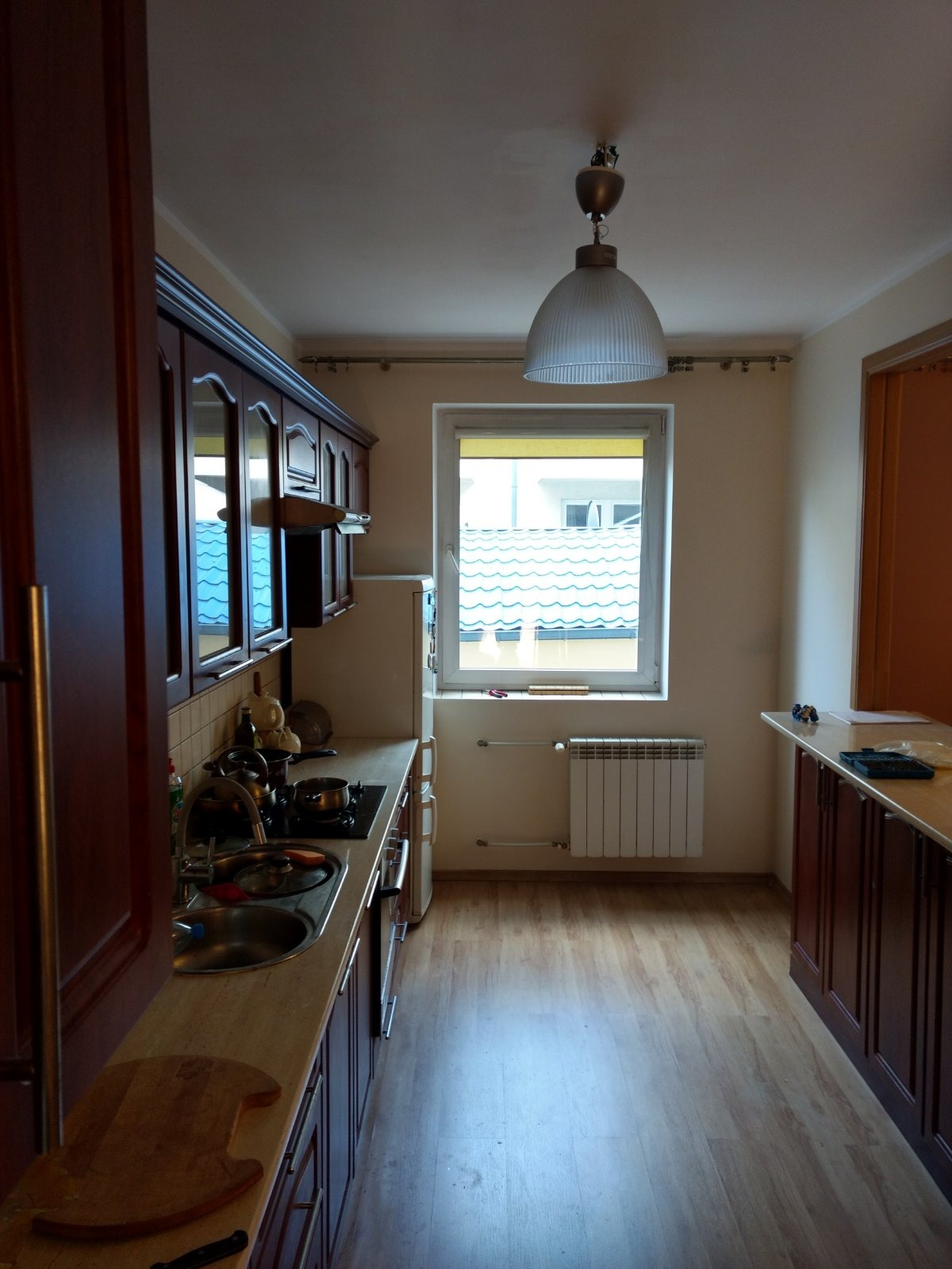 Kuchnia z ciemnymi, drewnianymi szafkami, blatem w odcieniach beżu i brązu oraz jasną podłogą z paneli; widoczne okno z widokiem na błękitny dach sąsiedniego budynku.