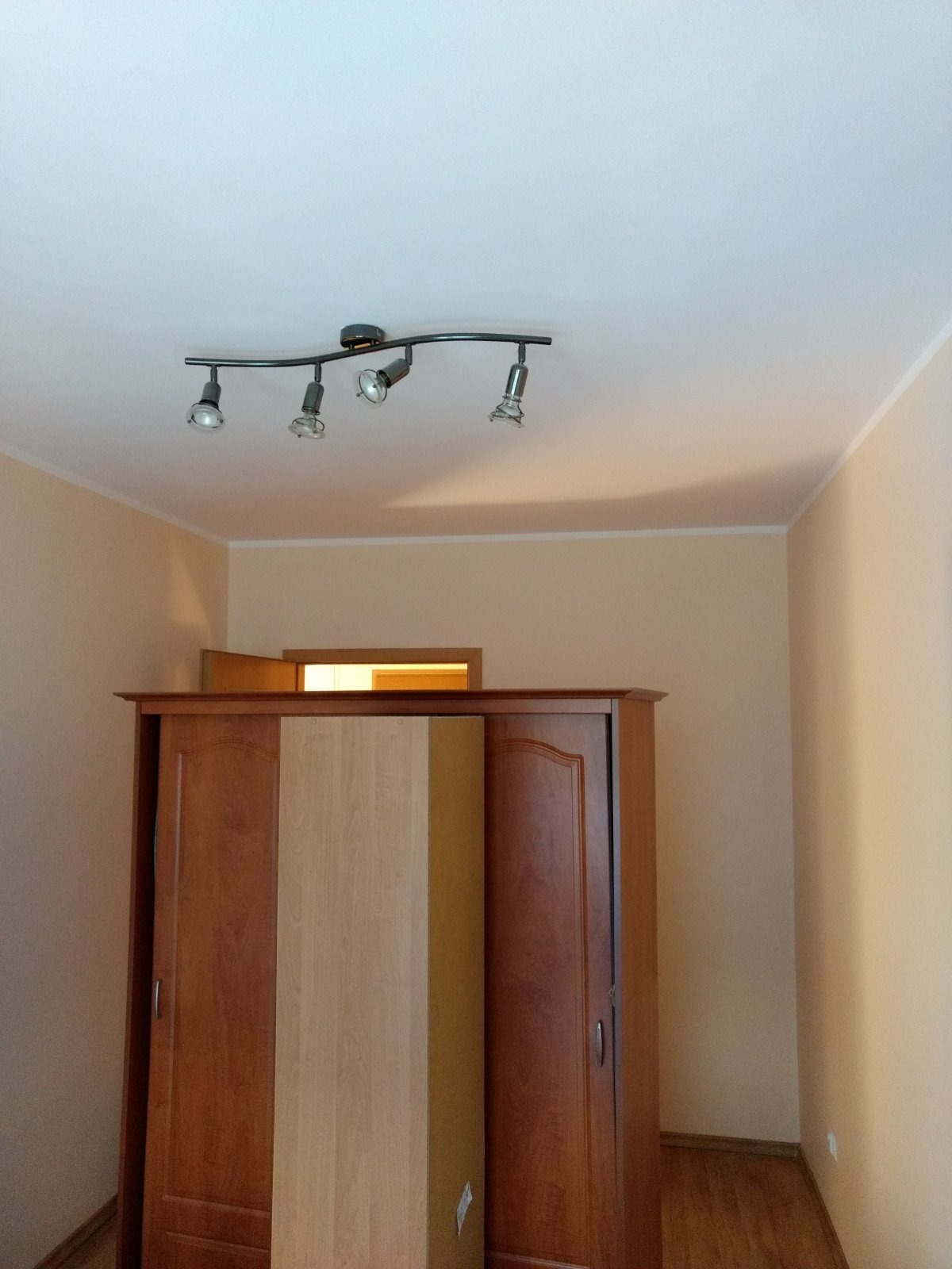 Wnętrze pokoju z beżowymi ścianami, białym sufitem i brązową szafą z przesuwnymi drzwiami, oświetlone listwą z czterema reflektorami.