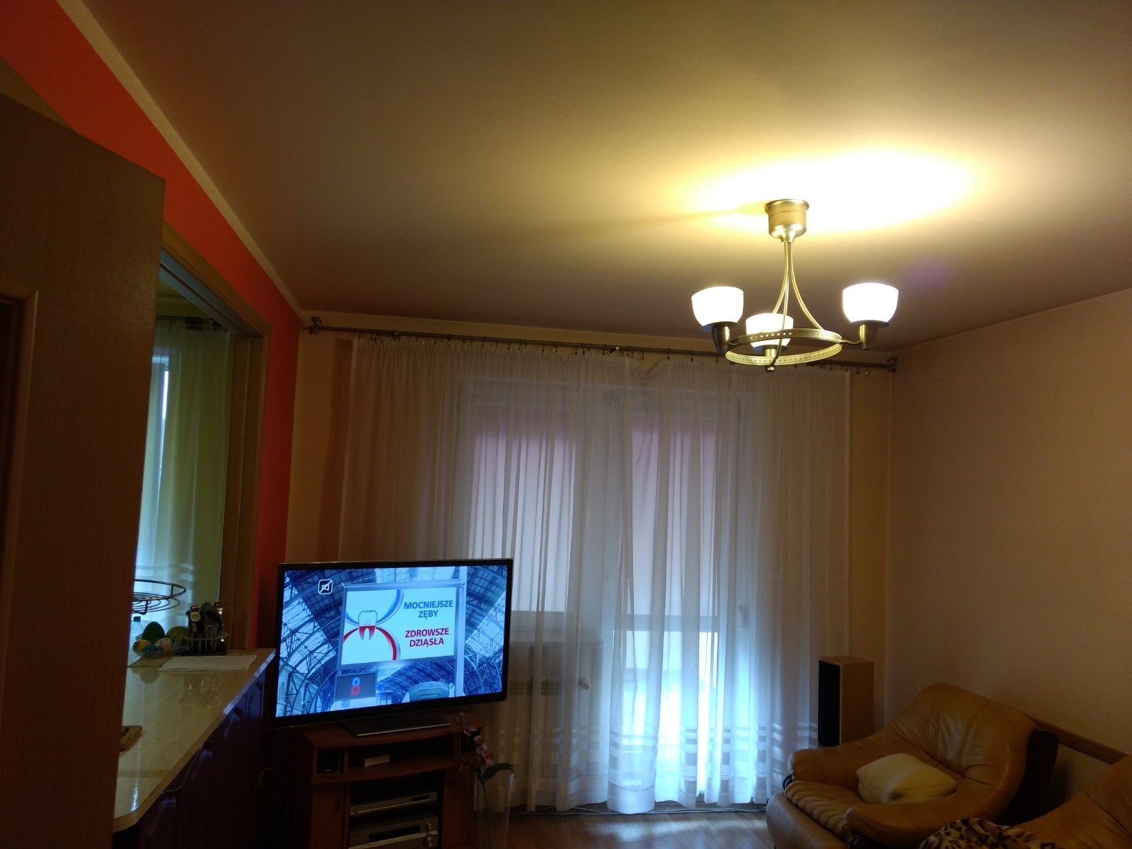 Jasne wnętrze pokoju z beżowymi ścianami, białymi zasłonami i brązową sofą, oświetlone przez żyrandol z trzema kloszami i lampą sufitową, z widocznym telewizorem wyświetlającym reklamę.