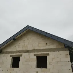 Surowy stan budynku z białych bloczków z zamontowanym, ciemnoszarym dachem blaszanym, widoczne otwory okienne i szary, pochmurny krajobraz w tle.