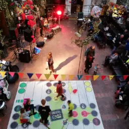 Widok z góry na podwórze z rozłożoną grą planszową, gdzie dzieci grają, w tle scena z zespołem muzycznym i publicznością.