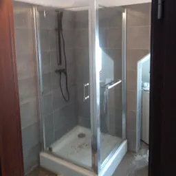 Szklana kabina prysznicowa z chromowanymi elementami i szarą armaturą na tle płytek w odcieniach szarości, widoczna pralka w tle.