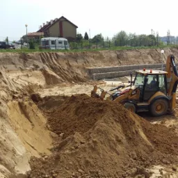 Żółta koparko-ładowarka CAT 432F podczas prac ziemnych na placu budowy, widoczny wykop, zabezpieczone gabionami skarpy i budynek w stanie surowym zamkniętym.