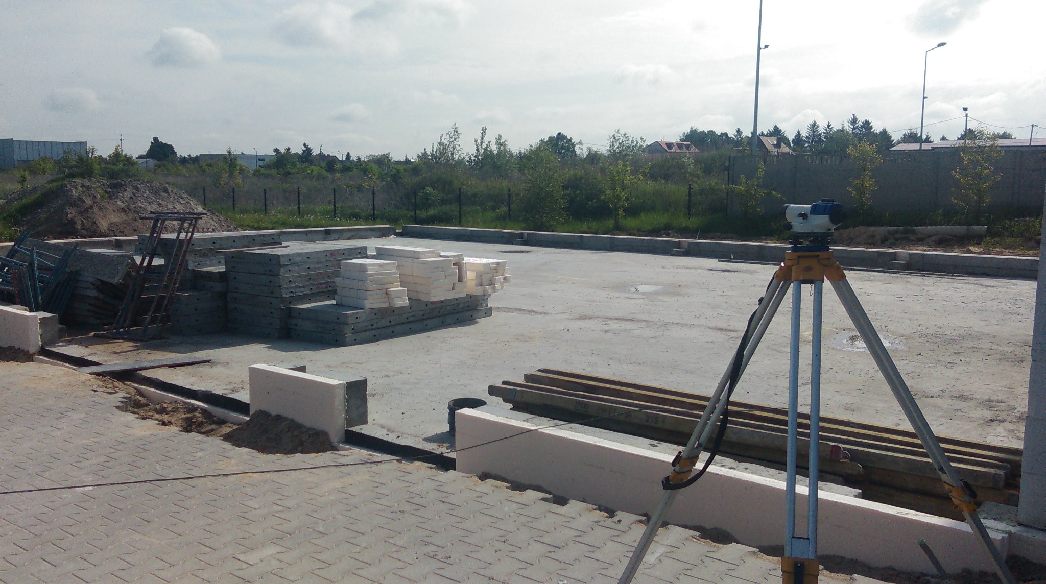 Budowa fundamentów: poziomica optyczna na trójnogu, ułożone bloczki betonowe i termoizolacyjne, przygotowany teren z widocznymi wykopami pod instalacje.