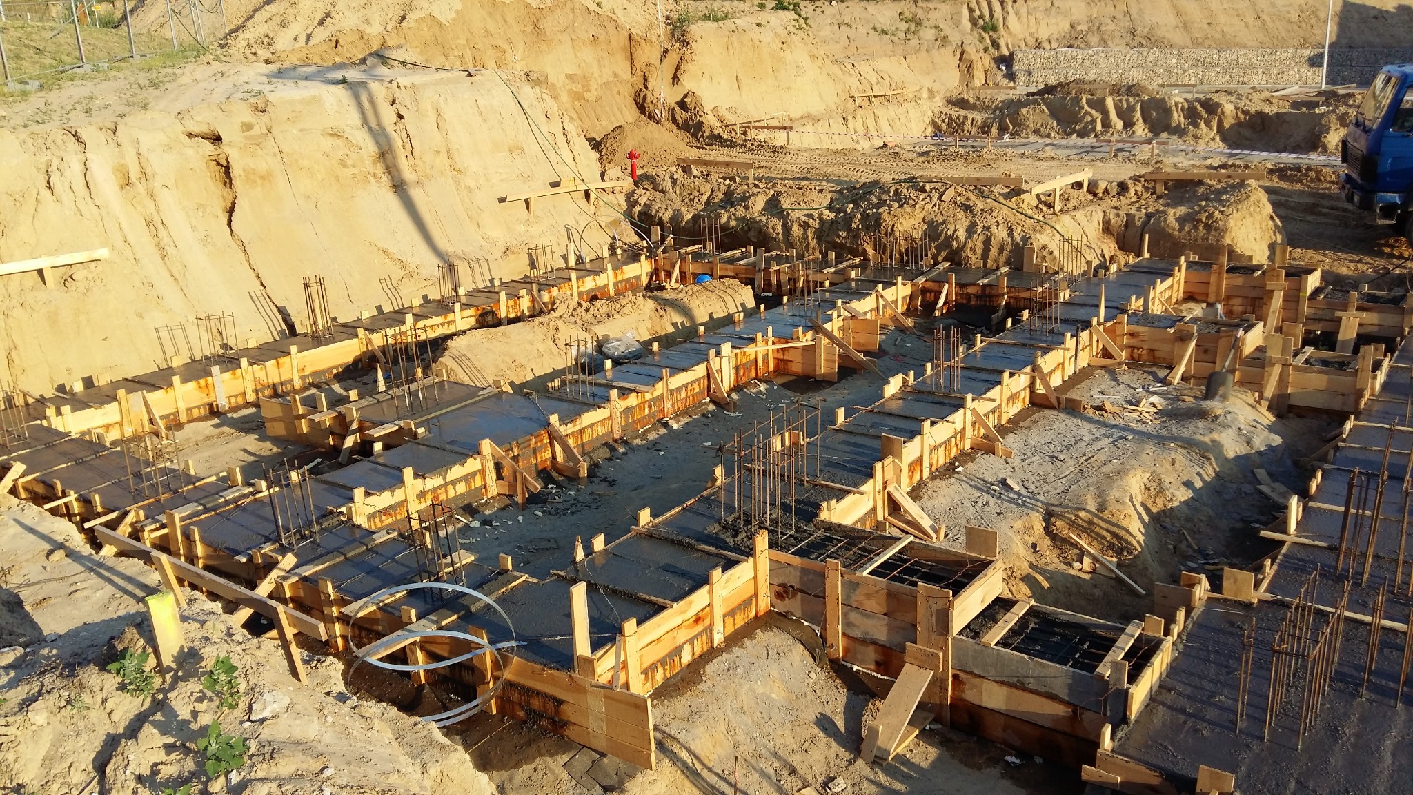 Wysoki kąt widzenia na fundamenty budynku w trakcie wylewania betonu, widoczne drewniane szalunki i wystające pręty zbrojeniowe, w tle wykop ziemny i częściowo widoczny niebieski samochód dostawczy.