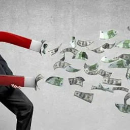 Mężczyzna w garniturze trzyma wielki, czerwony magnes podkowiasty, przyciągający strumień banknotów dolarowych i euro na tle szarej ściany.