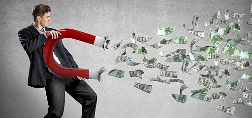 Mężczyzna w garniturze trzyma wielki, czerwony magnes podkowiasty, przyciągający strumień banknotów dolarowych i euro na tle szarej ściany.