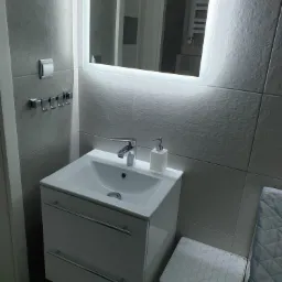 Nowoczesna łazienka z umywalką nablatową, szafką z dwiema szufladami, podświetlanym lustrem i szarymi płytkami.