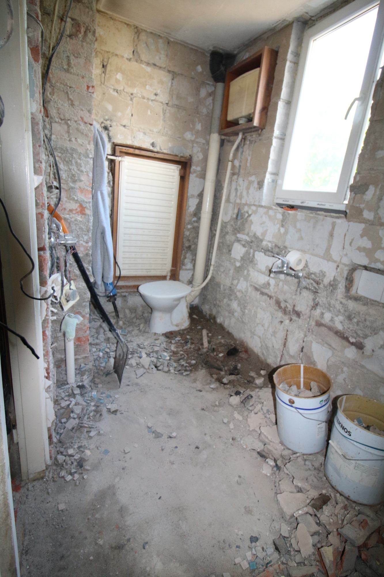 Łazienka w trakcie demontażu, widoczne fragmenty gruzu, odsłonięte cegły, stara toaleta, oraz narzędzia budowlane.