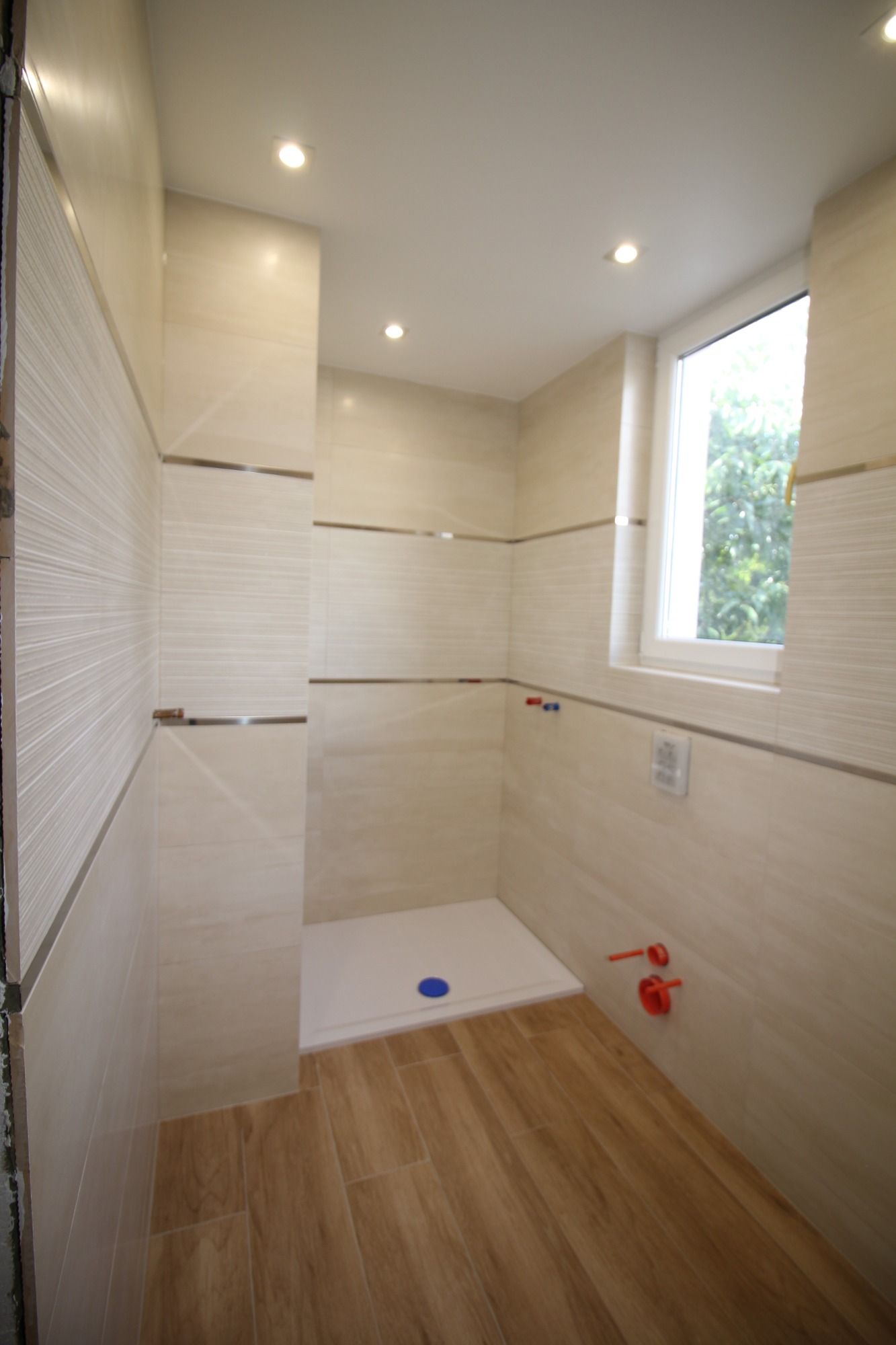 Jasna łazienka po remoncie z kabiną prysznicową typu walk-in, beżowe płytki ścienne z poziomymi paskami, drewnopodobna podłoga, okno z widokiem na zieleń.