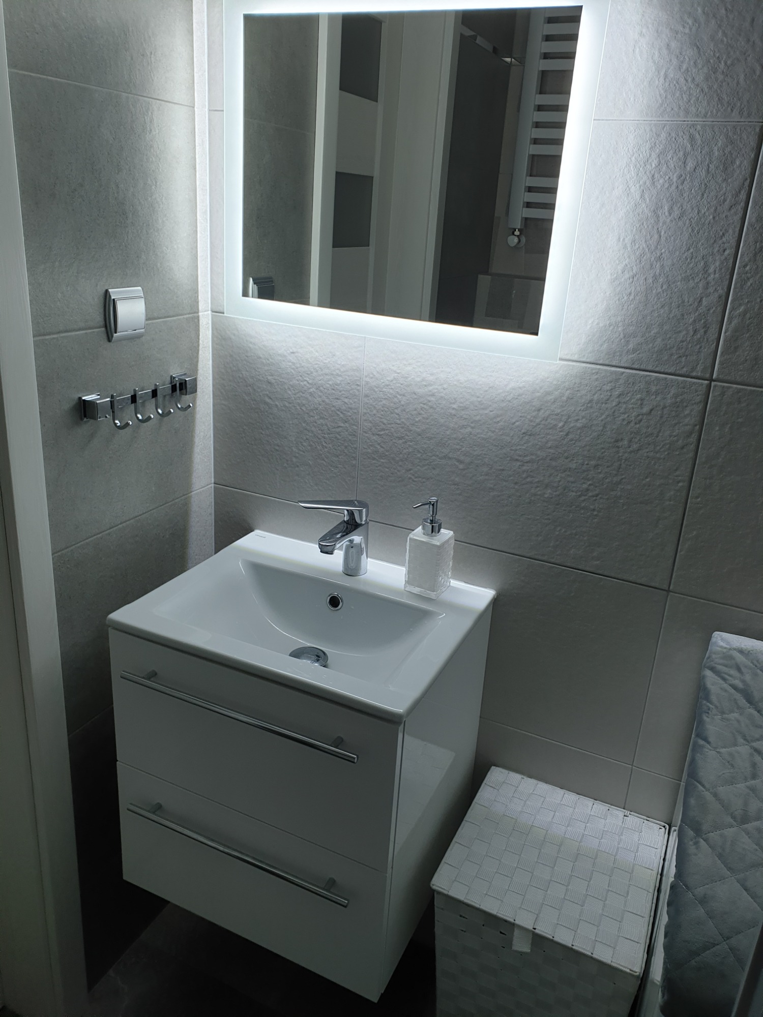 Nowoczesna łazienka z umywalką nablatową, szafką z dwiema szufladami, podświetlanym lustrem i szarymi płytkami.
