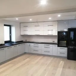 Nowoczesna kuchnia z białymi szafkami z czarnymi uchwytami, czarnym blatem i sprzętem AGD LG, wykończona jasną drewnianą podłogą.