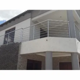 Balkon z nowoczesną balustradą z poziomych, cienkich prętów ze stali nierdzewnej, widok z dołu, szara kolumna podtrzymująca balkon, elewacja budynku w odcieniach bieli i szarości.