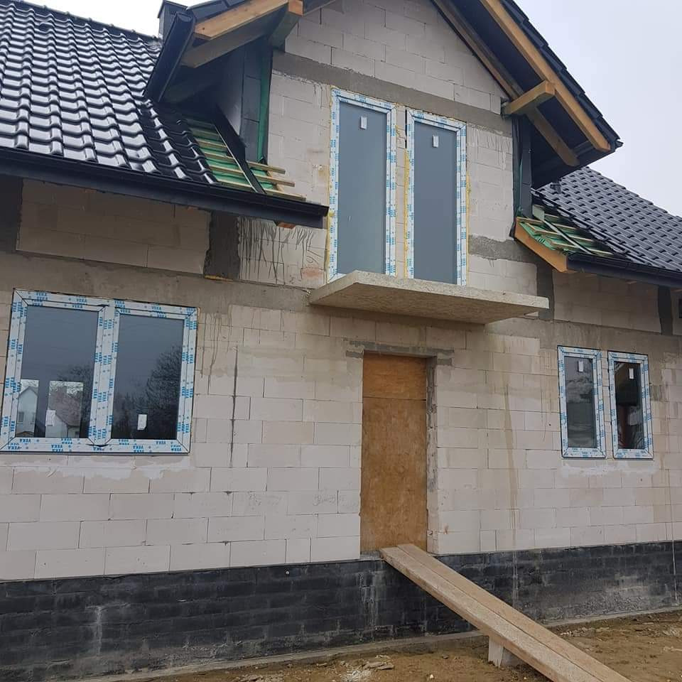 Surowy stan budynku jednorodzinnego z białego gazobetonu, z zamontowanymi oknami PCV z folią ochronną i tymczasowymi drzwiami z płyty, widoczny fragment dachu z czarną dachówką i deskowaniem.