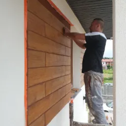 Pracownik na rusztowaniu nakłada zaprawę na elewację imitującą drewniane deski, zabezpieczoną pomarańczową taśmą malarską.