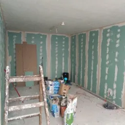 Wnętrze w trakcie montażu płyt kartonowo-gipsowych, widoczne szpachlowane łączenia, drabina malarska i worki z materiałami budowlanymi, częściowo zabudowana wnęka z półkami.