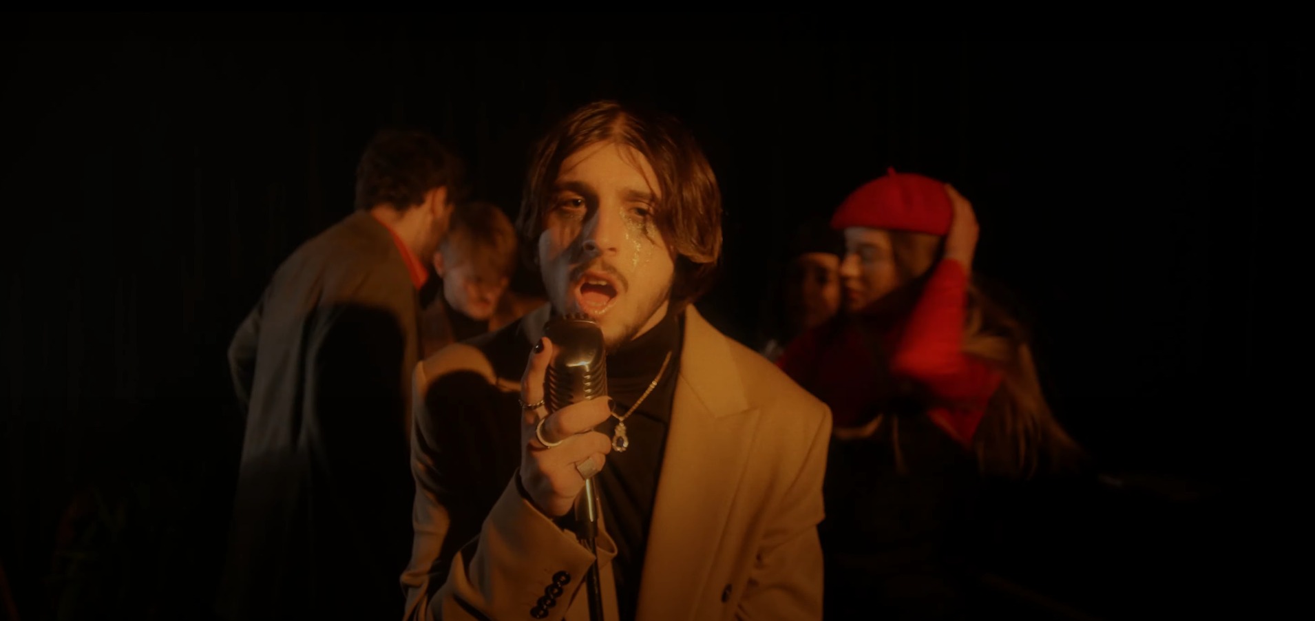 Młody mężczyzna z brokatem pod oczami, śpiewa do vintage mikrofonu na tle innych osób w ciemnym pomieszczeniu, prawdopodobnie podczas występu.