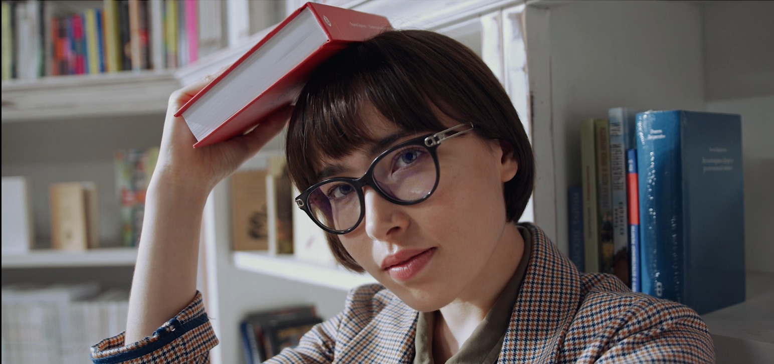 Kobieta w okularach trzymająca czerwoną książkę na głowie, półka z książkami w tle.