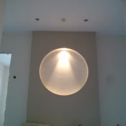 Niedokończone wnętrze z okrągłą lampą na ścianie, widoczne puszki farby Optiva, kartony i gniazdko elektryczne, surowy stan wykończenia.