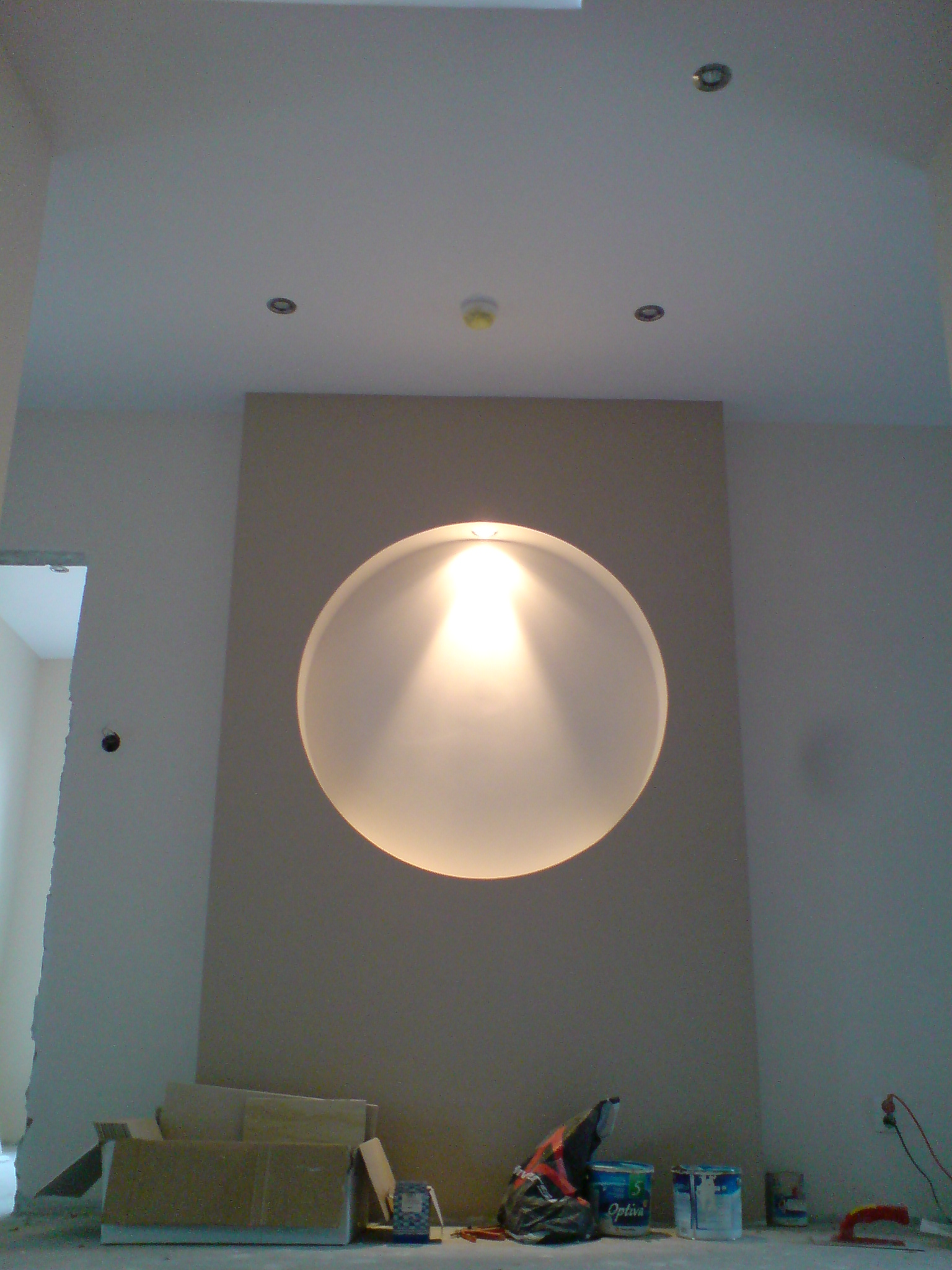 Niedokończone wnętrze z okrągłą lampą na ścianie, widoczne puszki farby Optiva, kartony i gniazdko elektryczne, surowy stan wykończenia.