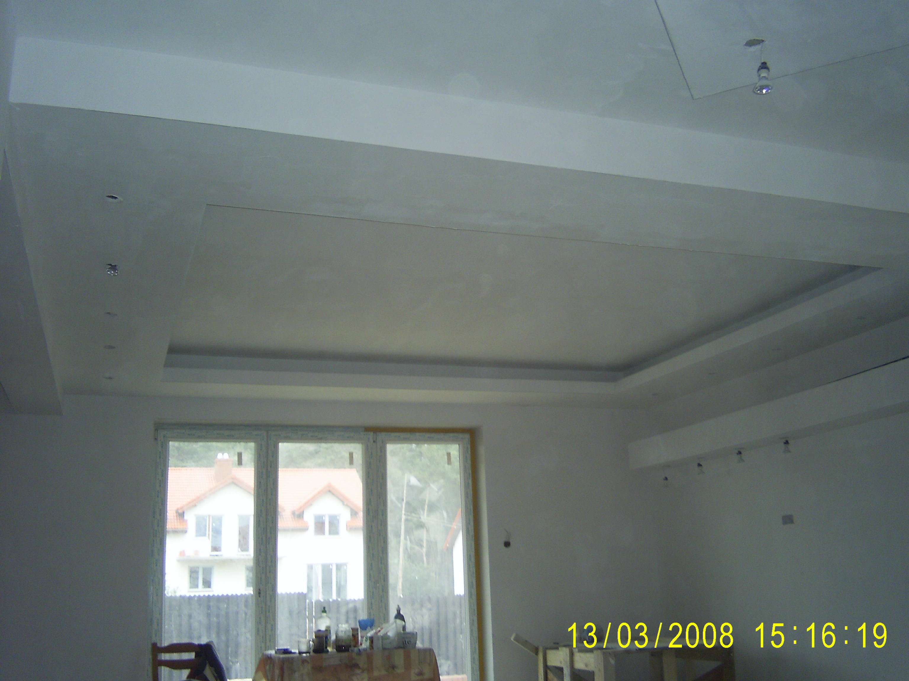 Sufit podwieszany z oświetleniem punktowym w trakcie prac wykończeniowych, widoczne okno z widokiem na dom i elementy wyposażenia wnętrza.