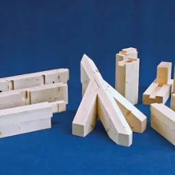 Elementy drewniane z wyfrezowanymi otworami i połączeniami ciesielskimi, przygotowane do montażu konstrukcji dachowej na tle niebieskiego tła.