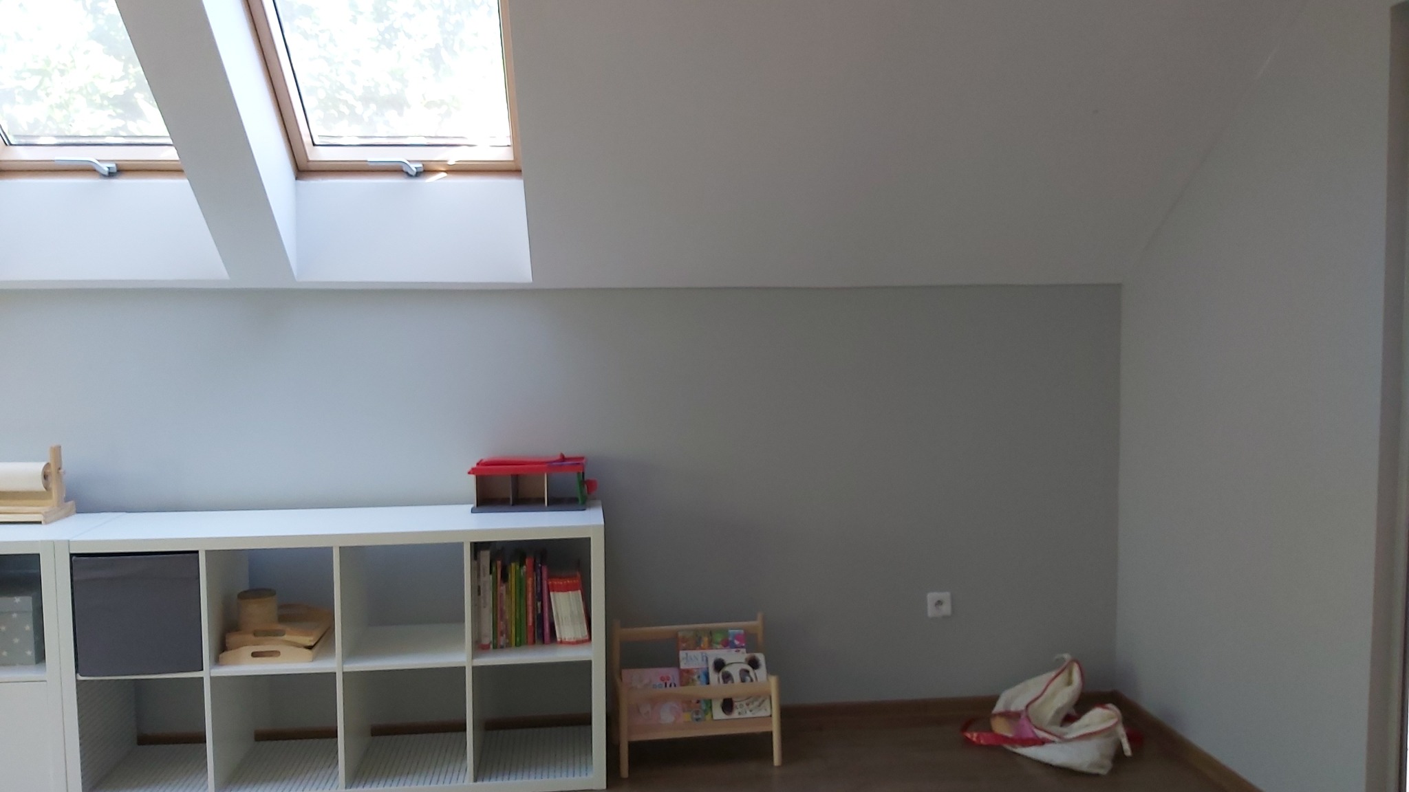 Poddasze z oknem dachowym, pomalowane na szaro, z białym regałem na zabawki i książki. Na regale widoczne pudełka, książki i zabawki. Na podłodze mały regał z książkami i torba.