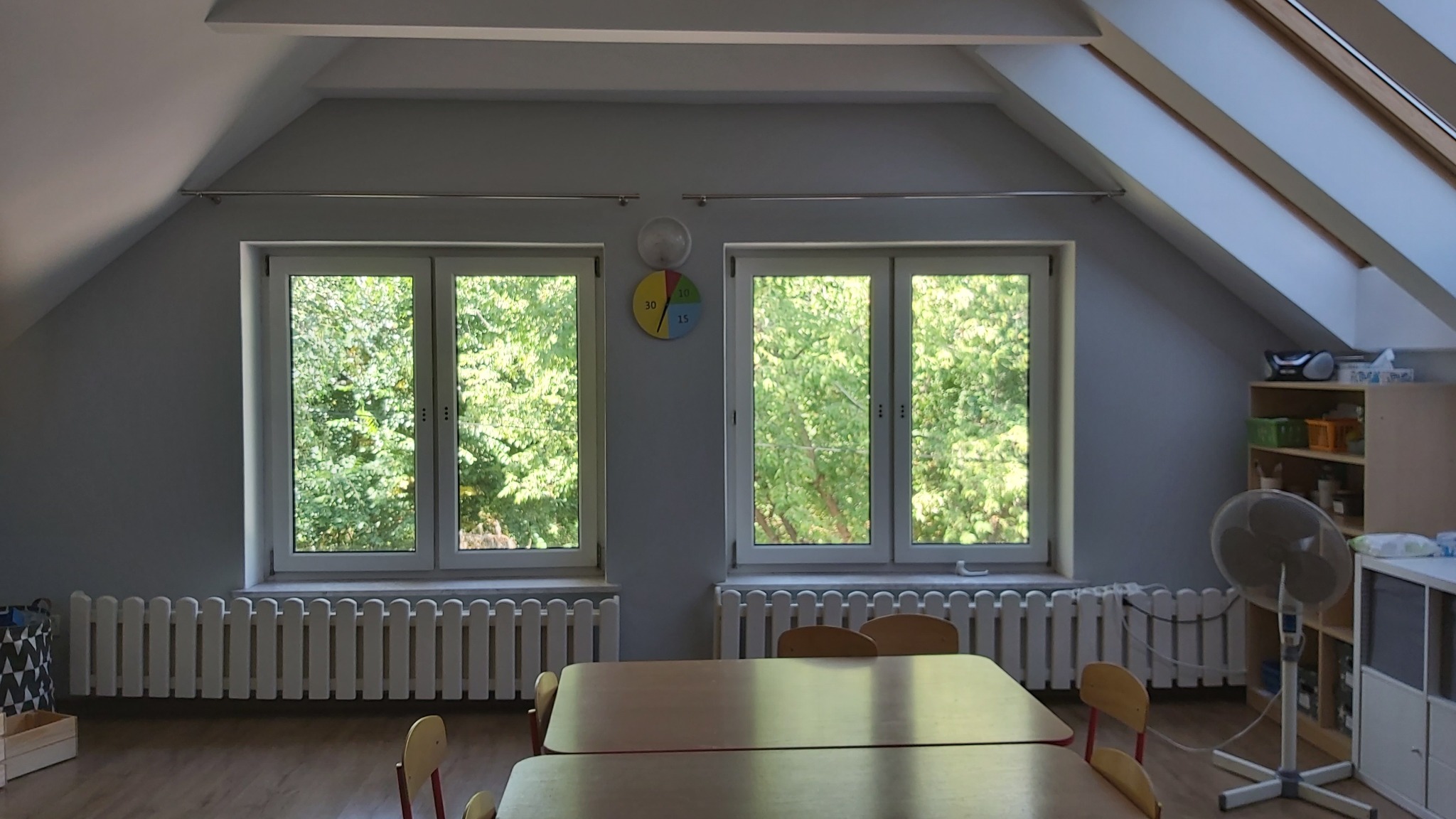 Pokój na poddaszu z dwoma oknami z widokiem na zieleń, zegar na ścianie, meble i stoły dla dzieci, grzejniki dekoracyjne.