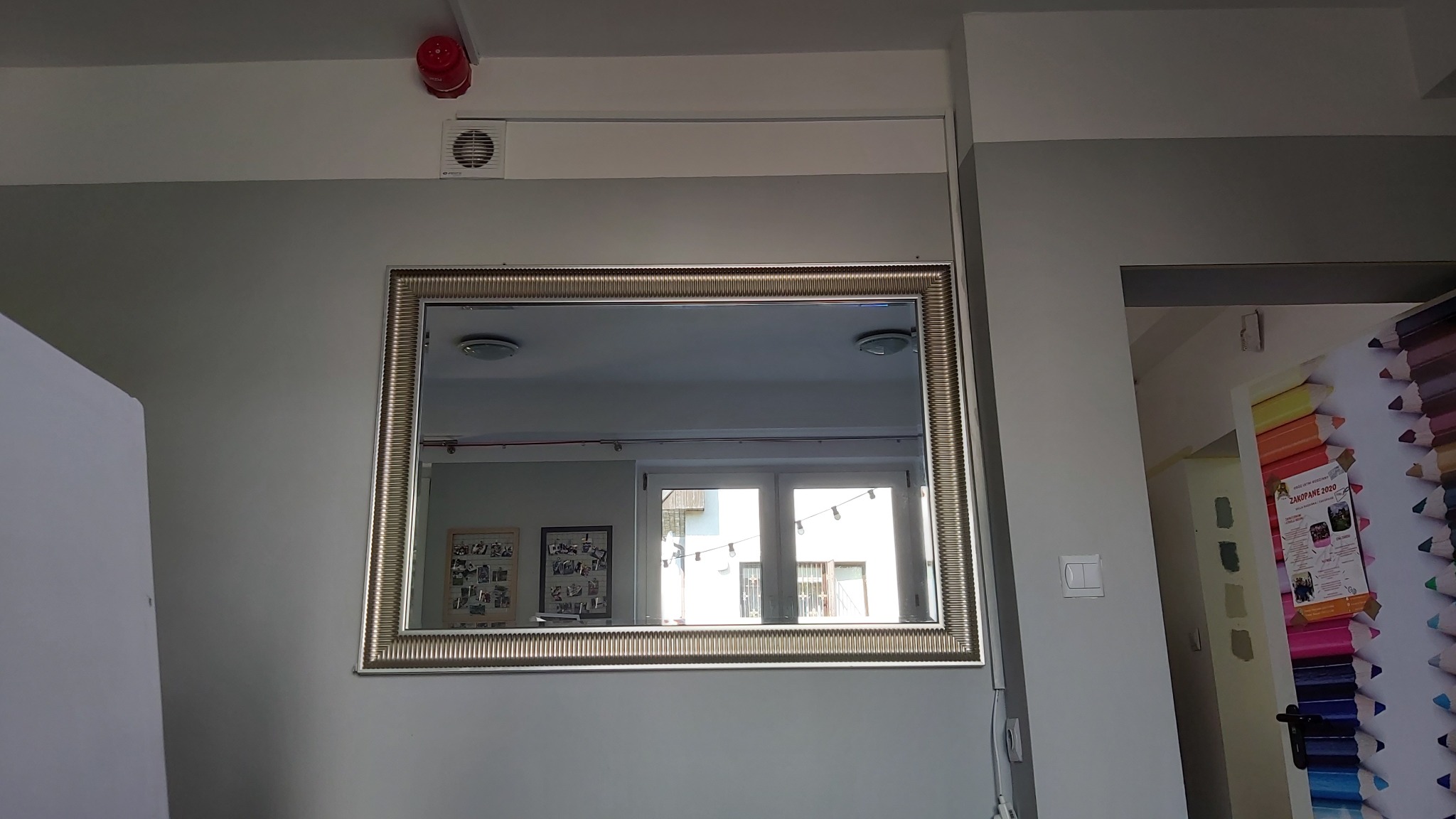 Lustro w złotej ramie zawieszone na szarej ścianie w odnowionym pomieszczeniu, z widocznym odbiciem okna i plakatów w tle.