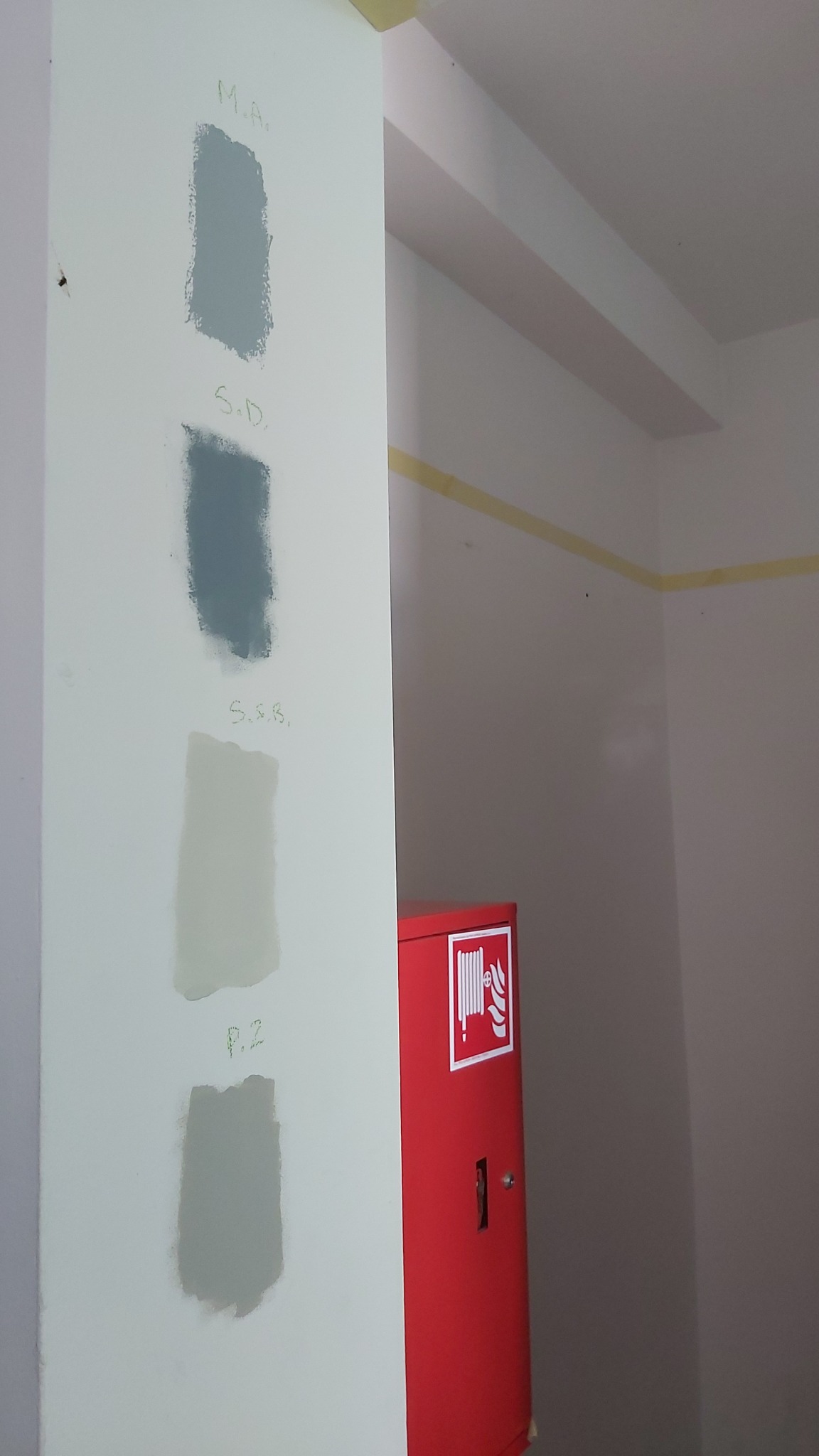 Ściana z próbkami farb w odcieniach szarości i beżu, z ręcznie napisanymi oznaczeniami nad każdą próbką, obok czerwona szafka hydrantowa.