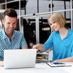 Mężczyzna i kobieta pochyleni nad białym laptopem na jasnym biurku, analizujący dane; w tle przeszklona ściana biurowa.