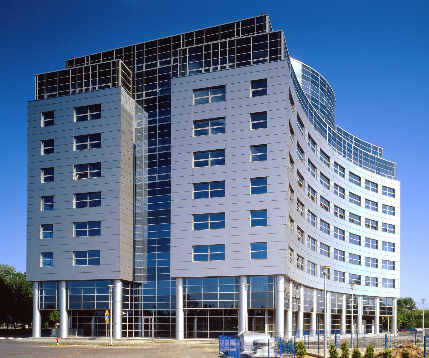 Nowoczesny budynek biurowy o srebrnej elewacji z wyraźnymi liniami i zakrzywioną szklaną fasadą, widoczny z poziomu ulicy w słoneczny dzień.