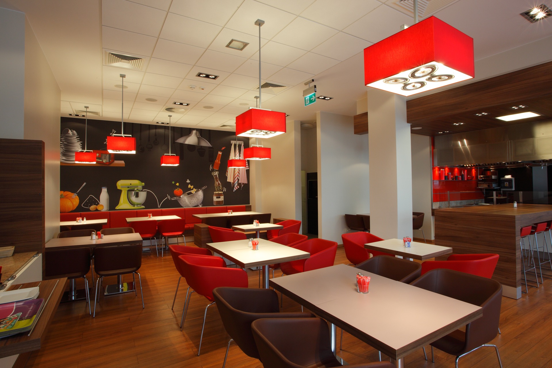 Wnętrze nowoczesnej restauracji z drewnianą podłogą, białymi stolikami i czerwonymi krzesłami, oświetlone czerwonymi lampami. Na ścianie czarny mural z motywami kulinarnymi, w tle widoczna otwarta...