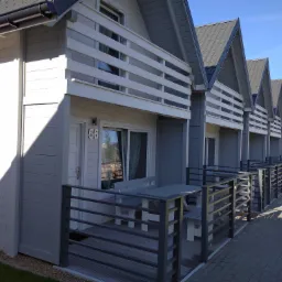 Szereg identycznych, szarych domków letniskowych z białymi balkonami i numerami, ustawionych wzdłuż brukowanej ścieżki.