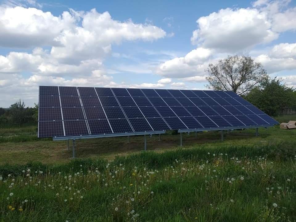Panel słoneczny zamontowany na metalowej konstrukcji, otoczony zieloną łąką z dmuchawcami, na tle błękitnego nieba z białymi obłokami.